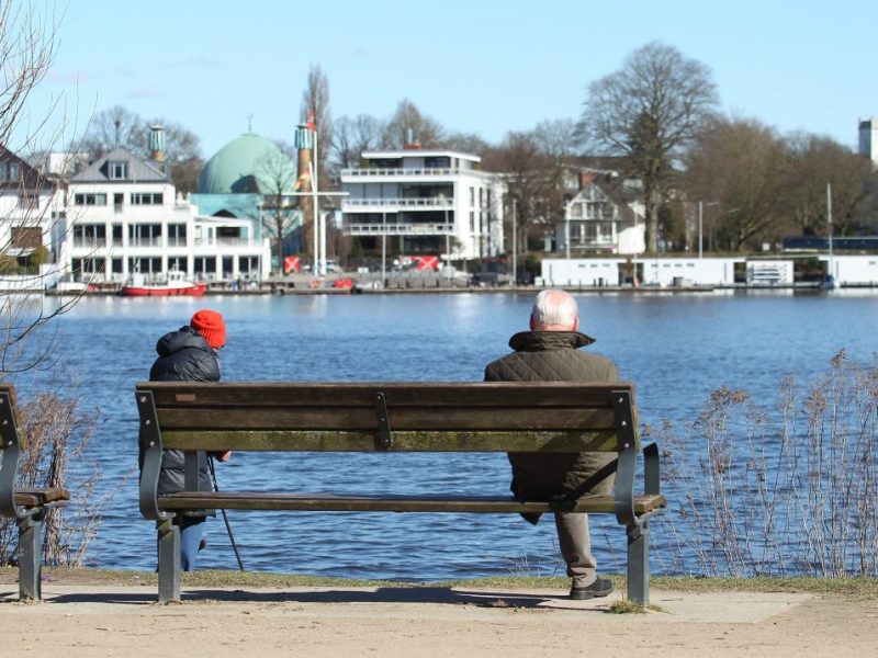 Ein Mann sitzt bei frühlingshaften Temperaturen und Sonnenschein auf einer Sitzbank an der Außenalster, Symbolbild